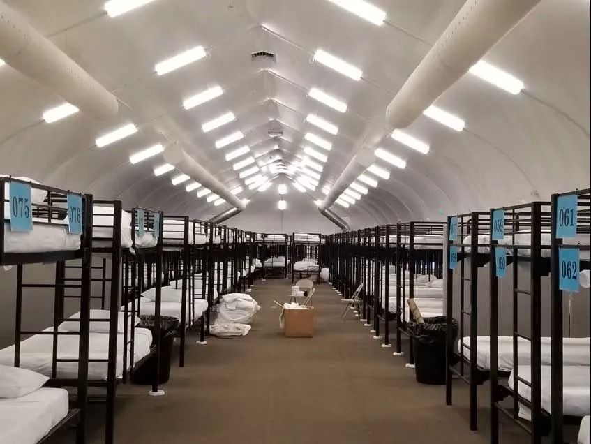 Interior view of a large temporary shelter with rows of bunk beds, lit by overhead lights.