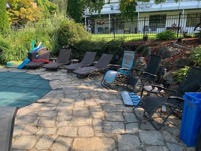 Patio with several lounge chairs and blue poolside seating beside greenery and a fence