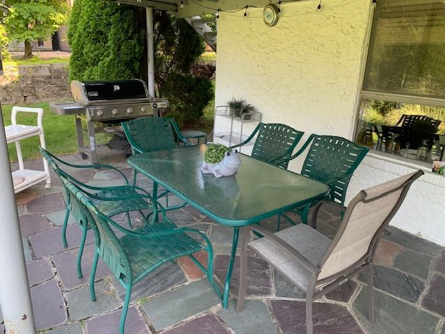 Patio dining area with green metal table and chairs beside a house, with a grill in the background