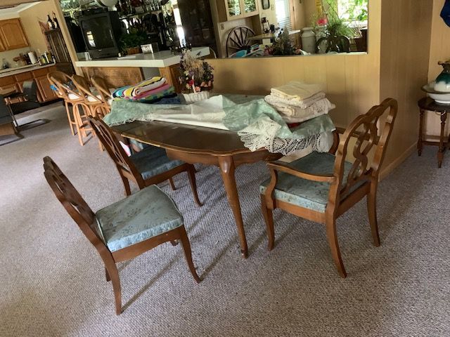 Wooden dining table with chairs in a living room, covered by a sheet and papers, near a kitchen pass-through.