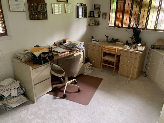 Cluttered home office with desk, chair, papers, and wood furniture by a window and blinds.