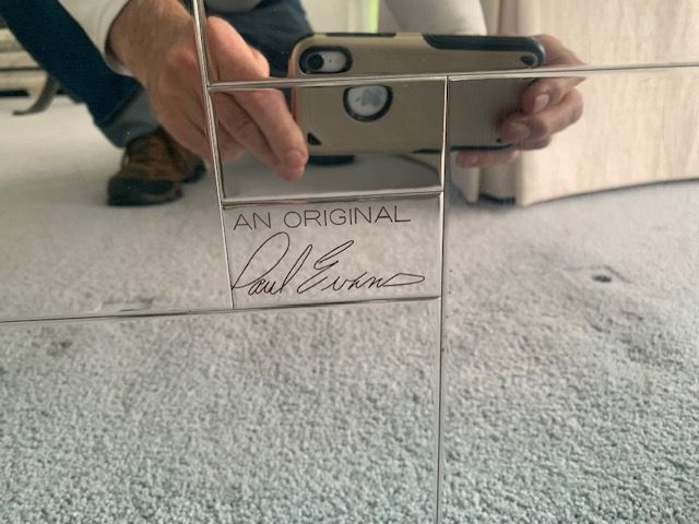 Phone behind a clear acrylic stand with “AN ORIGINAL” and a signature on a white surface