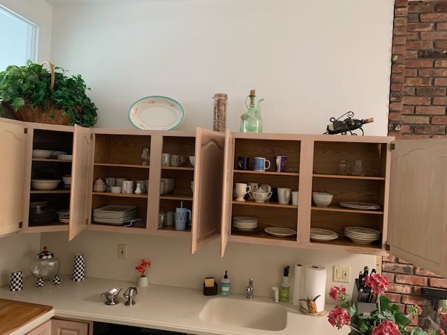 Kitchen with open wooden cabinets, dishes, and plants beside a brick fireplace.