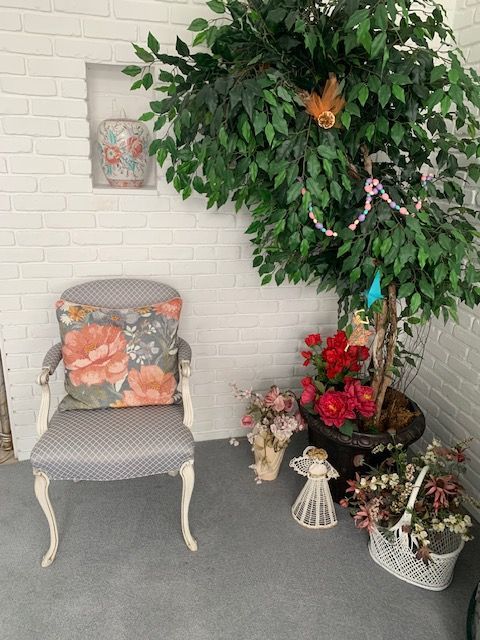Patio corner with floral chair, large potted tree, and red-and-white flower arrangements against a white brick wall