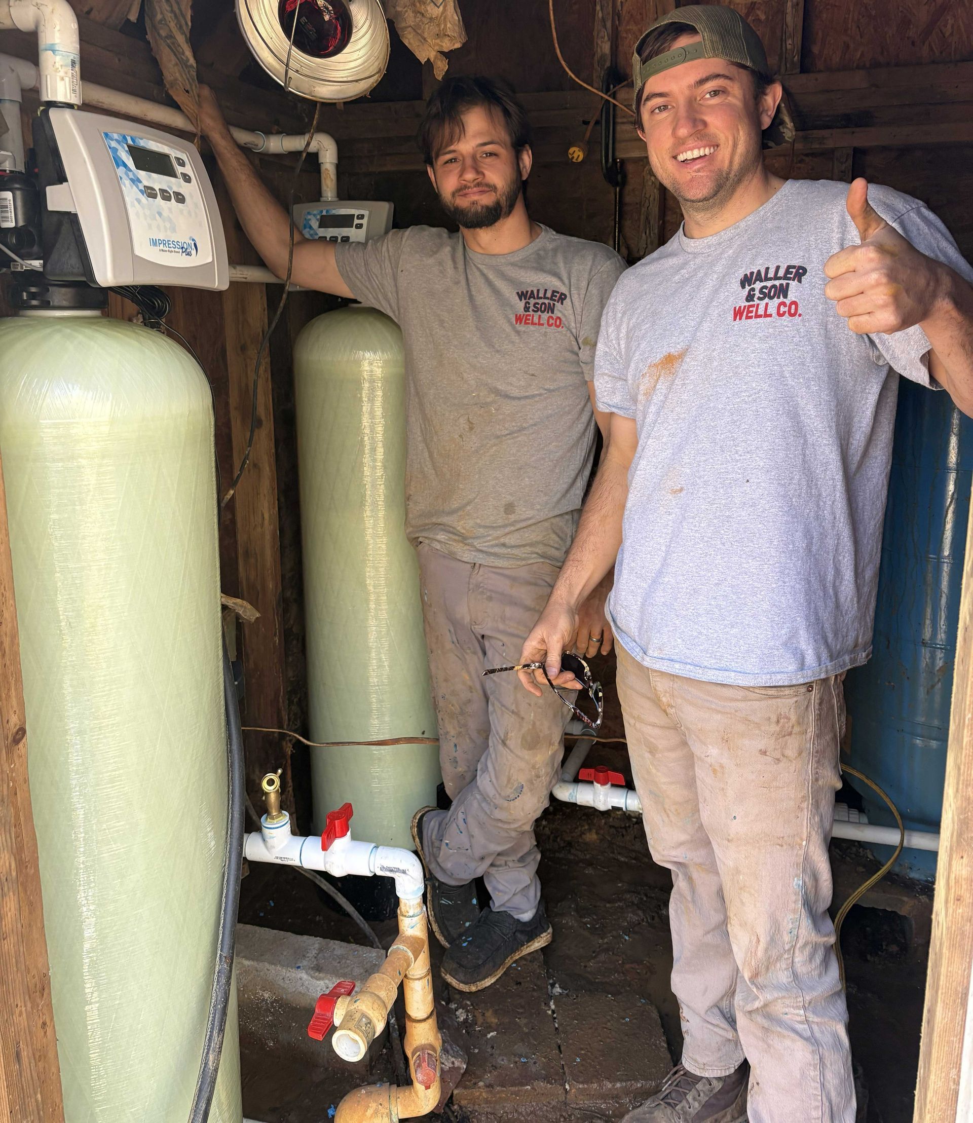 Two people in gray work shirts stand in a utility room with two large water filtration tanks, one giving a thumbs-up.