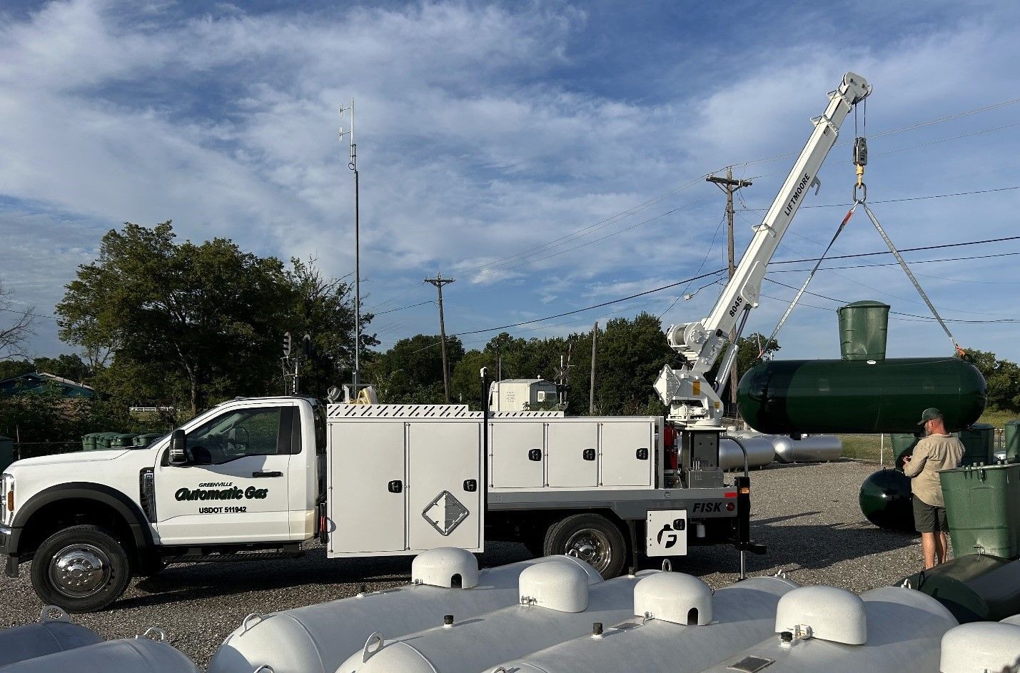 White truck with crane lifting a green propane tank; worker oversees the installation, surrounded by other tanks.