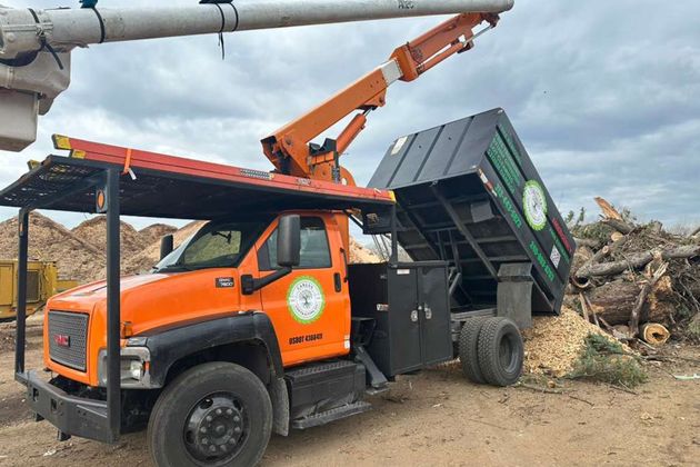 An orange GMC bucket truck with a lifted dump bed tilted to empty wood chips onto a pile at an outdoor site.