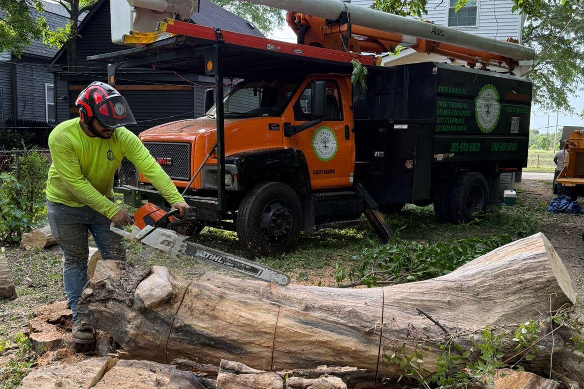 A worker in a high-visibility lime shirt and safety gear cuts a large log with a chainsaw in front of an orange tree truck.