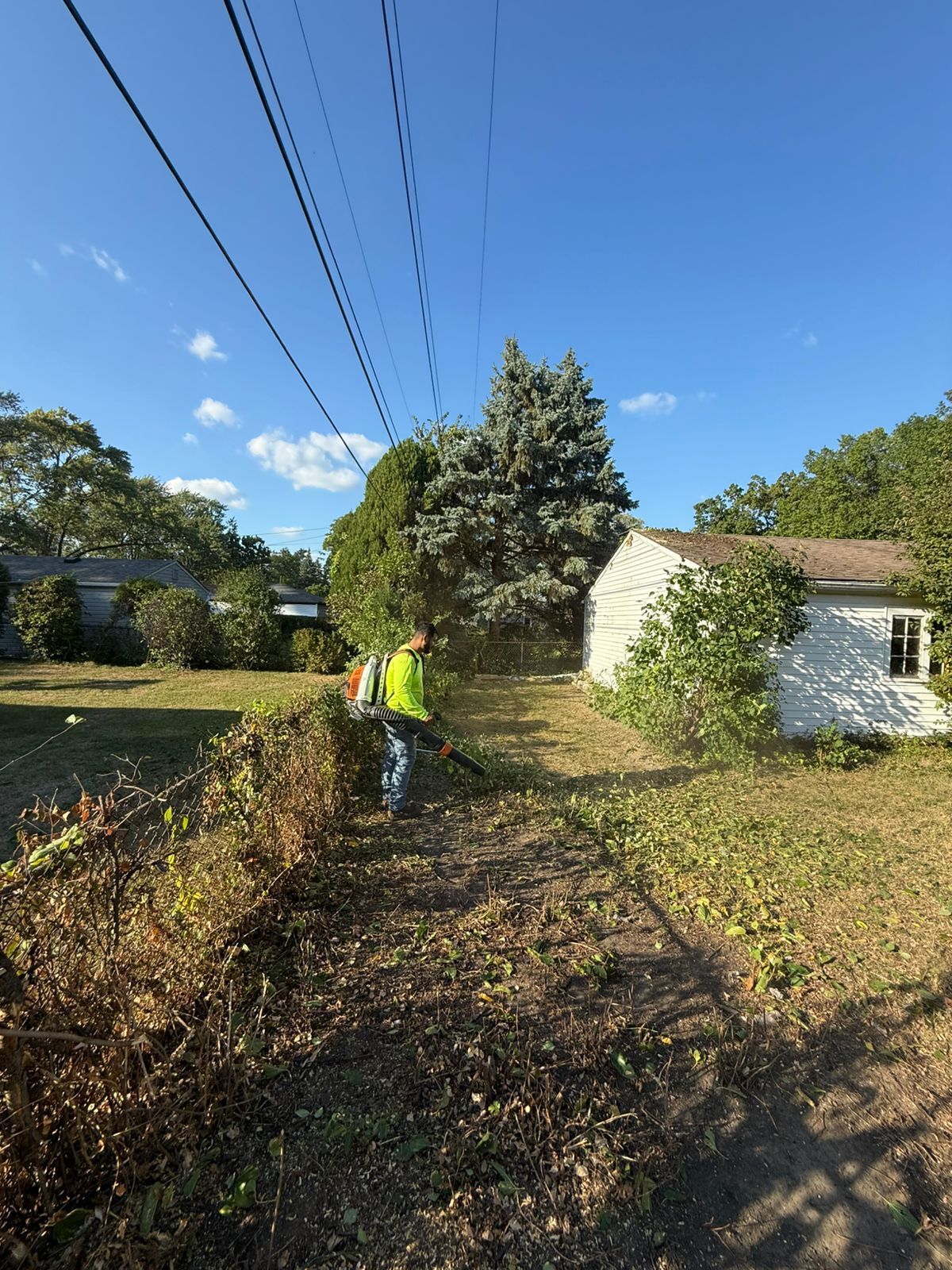 A person in a neon yellow shirt uses a leaf blower to clear debris near a chain-link fence and a white house.