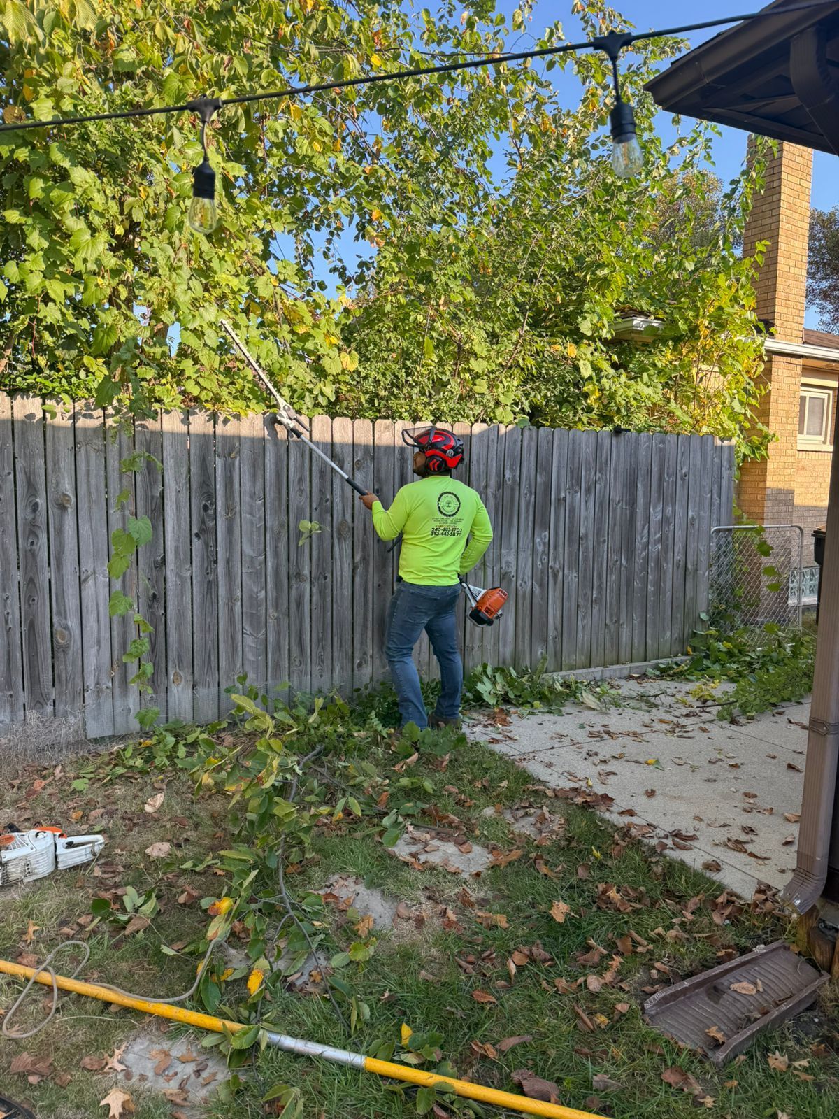 A worker in a neon yellow shirt and red helmet uses a pole saw to trim tree branches near a wooden fence.