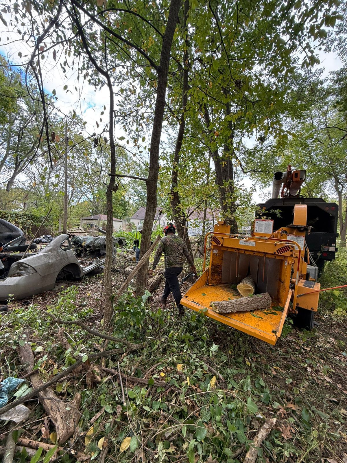A person walks past a bright yellow wood chipper in a wooded area near a parked car.