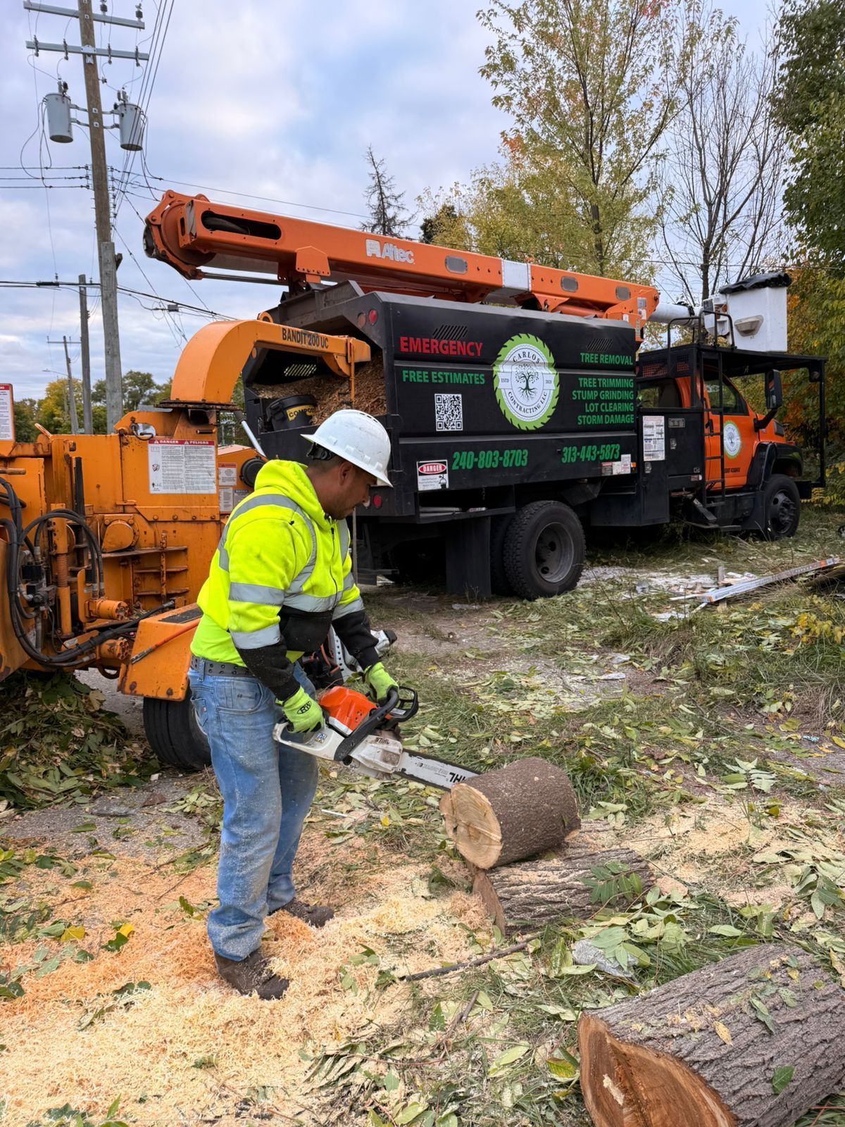 A worker in a high-visibility jacket uses a chainsaw to cut a log near a woodchipper truck at a job site.