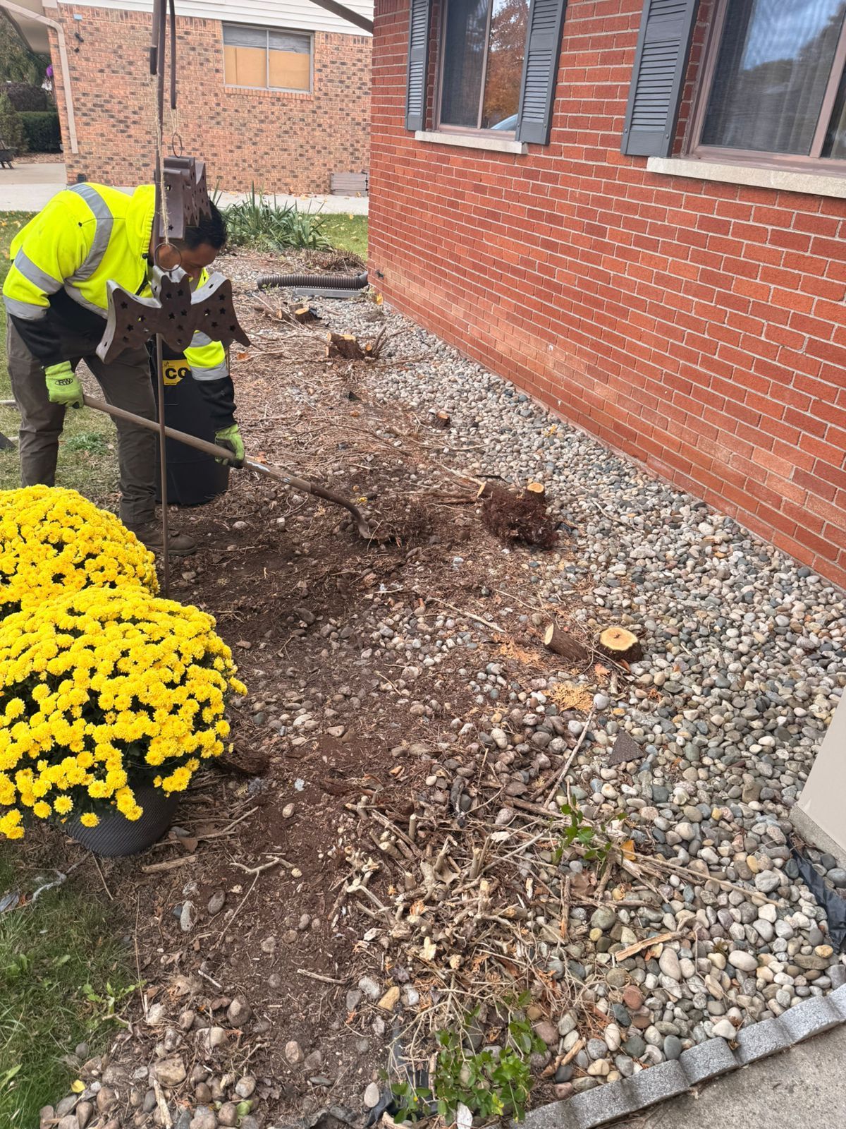 A worker in a high-visibility jacket uses a tool to clear debris from a rocky garden bed by a brick house.