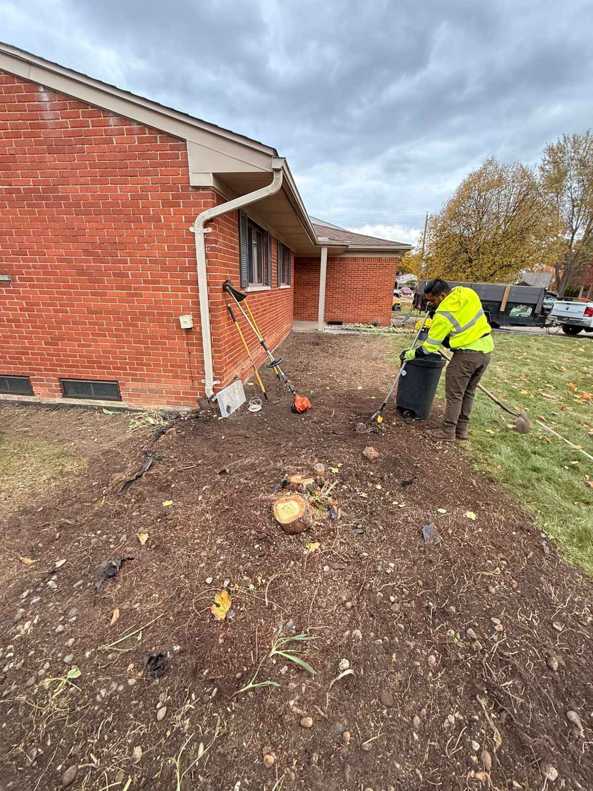 A person in a high-visibility jacket clears debris from a landscaped yard beside a brick house.