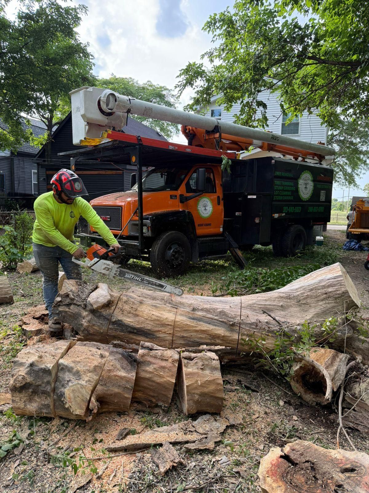 A worker in a high-visibility lime shirt cuts a large fallen log with a chainsaw near an orange utility truck.