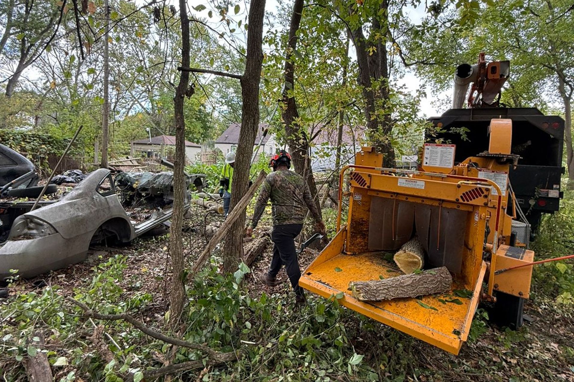 A person in protective gear operates a wood chipper near a wrecked car in a wooded area.