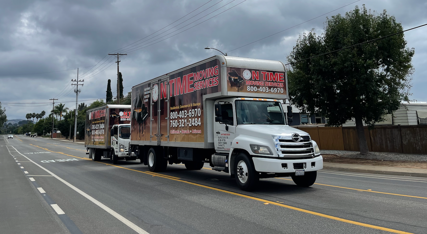 White moving truck pulling a smaller trailer on a road under a cloudy sky.