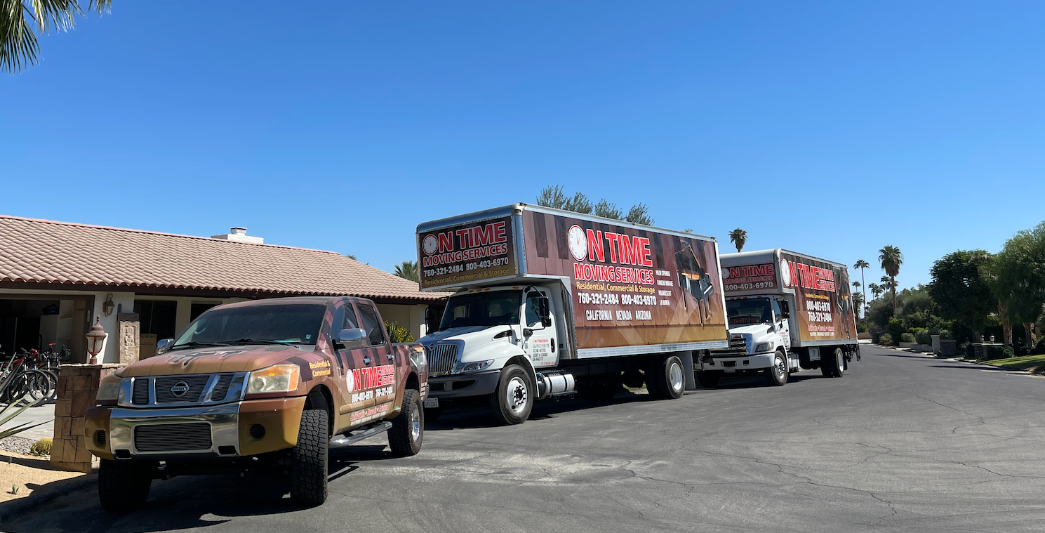 A moving company's trucks and a pickup truck parked on a residential street on a sunny day.