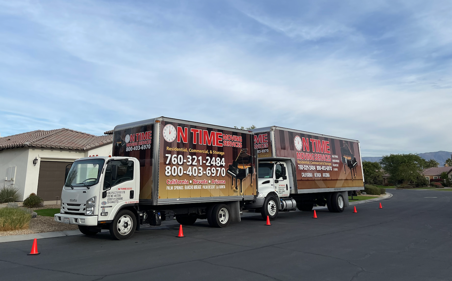 Two moving trucks parked on a street; one truck is pulling a trailer. The trucks are white with colorful advertisement wraps.