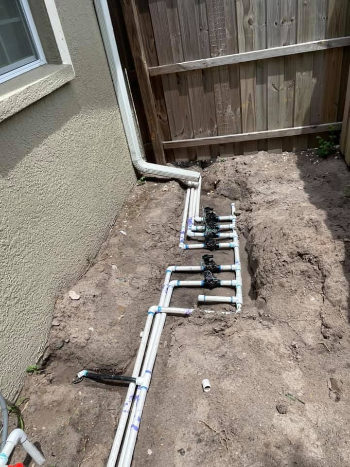 Irrigation system plumbing installed in a sandy trench beside a house and wooden fence.