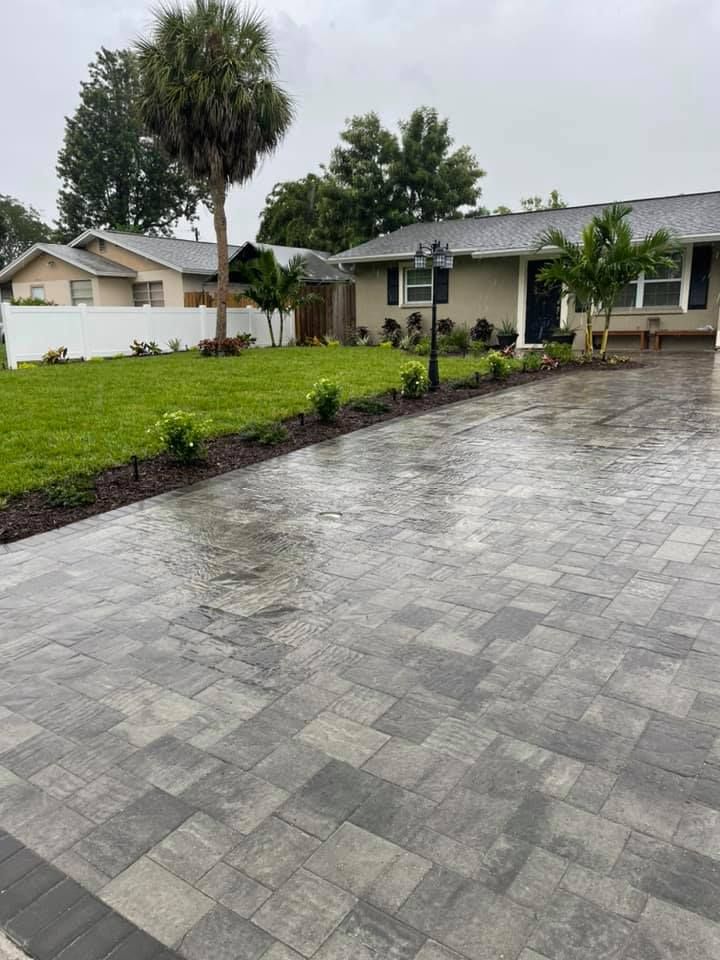 Brick paver driveway leads to a beige house with a manicured lawn and landscaping.
