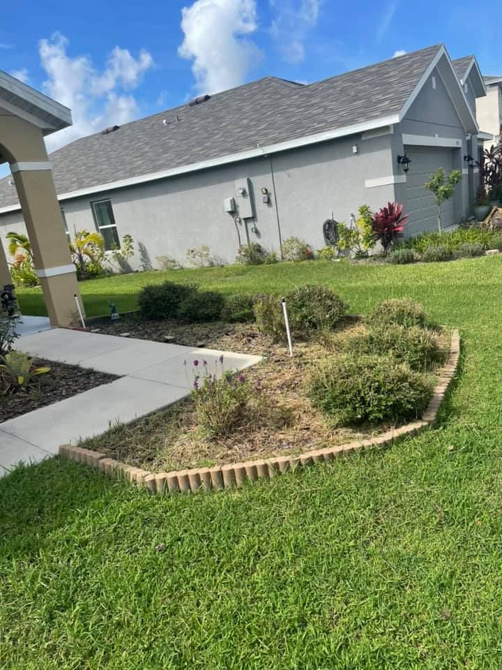 A flowerbed in front of a gray house with a concrete walkway on a sunny day.