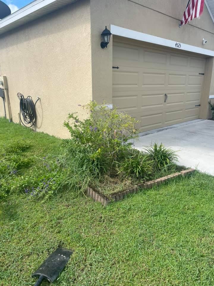 Tan house with a garage, landscaping, and a hose reel on the wall. The yard has green grass.