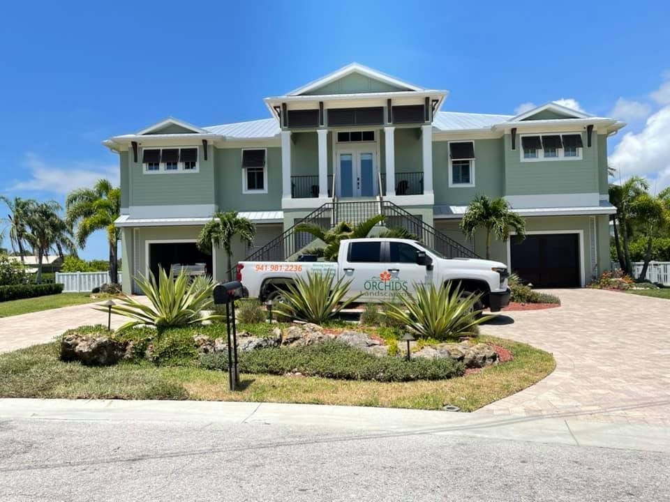 Two-story coastal house with light green siding and a white truck in the driveway. Landscaping includes green plants and rocks.
