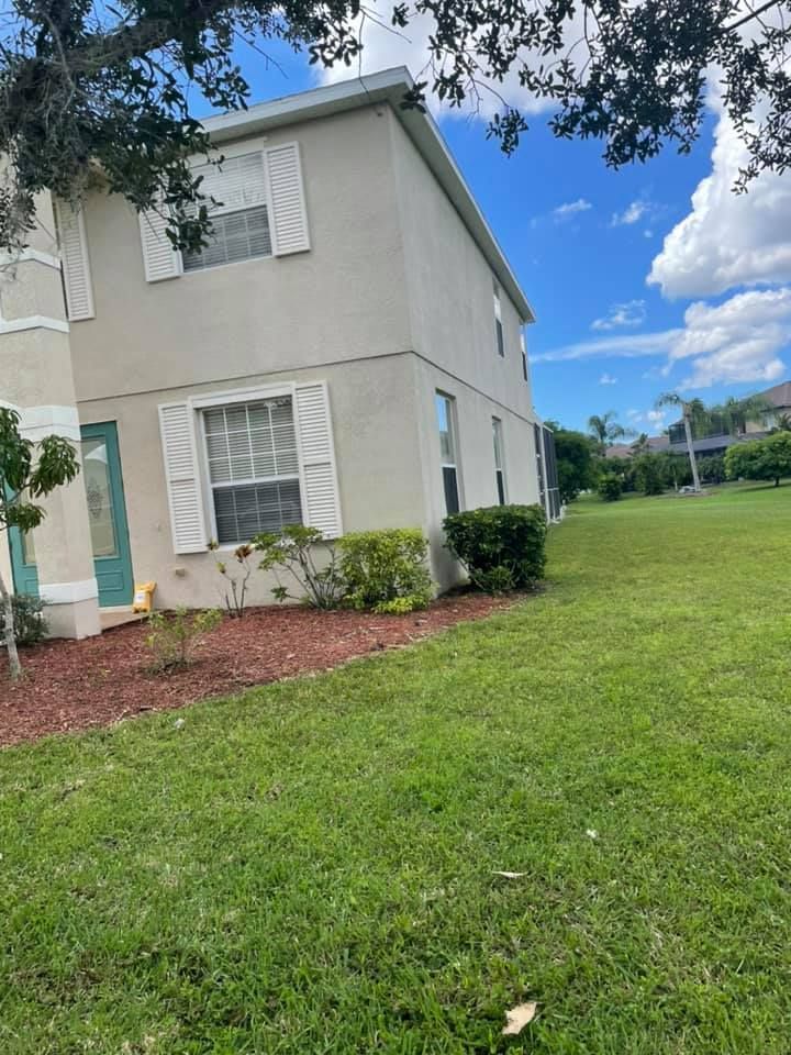 Two-story beige house with white shutters, teal door, and green lawn under a blue sky.