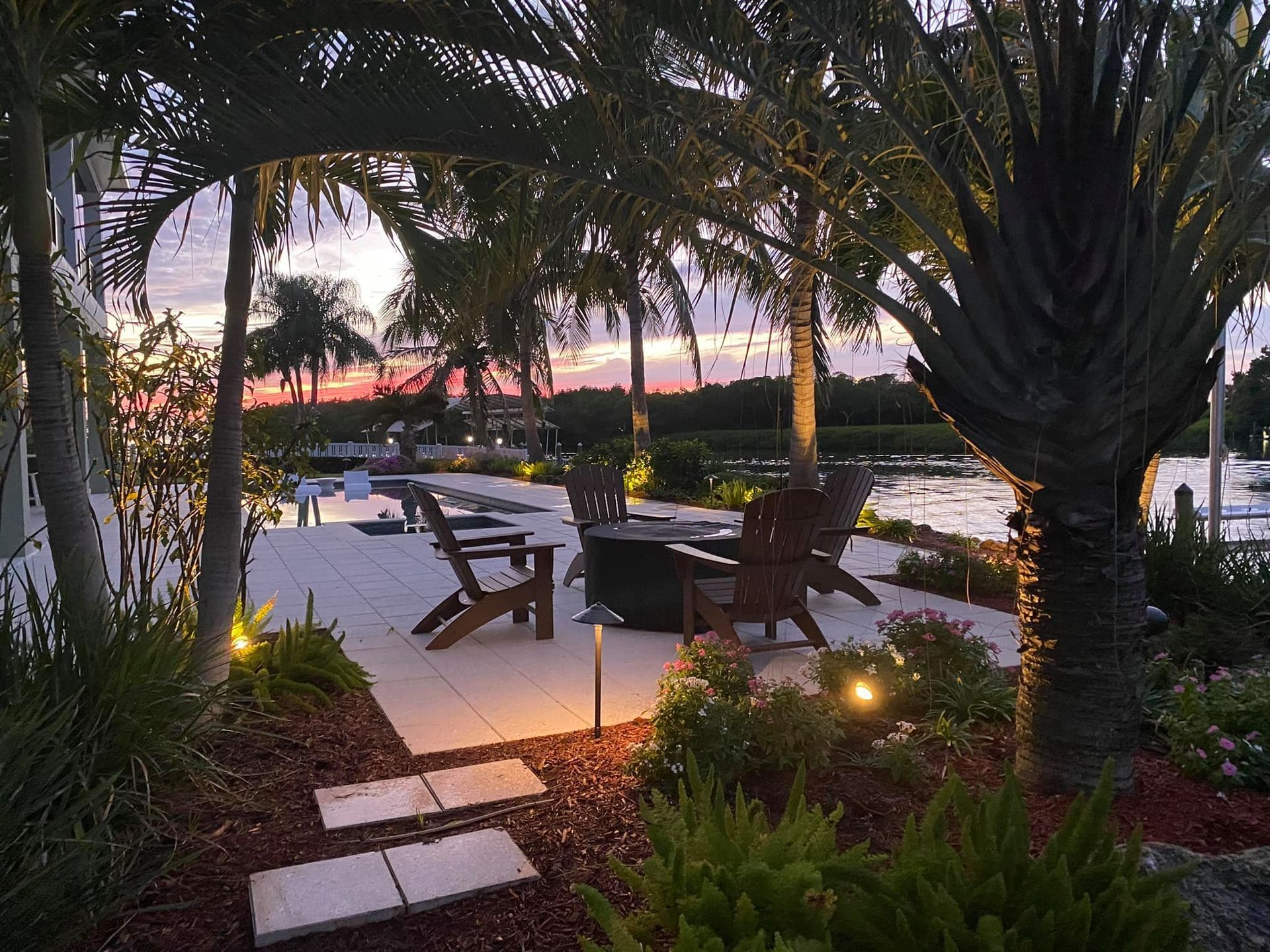 Patio with Adirondack chairs, fire pit, and palm trees at dusk overlooking water.