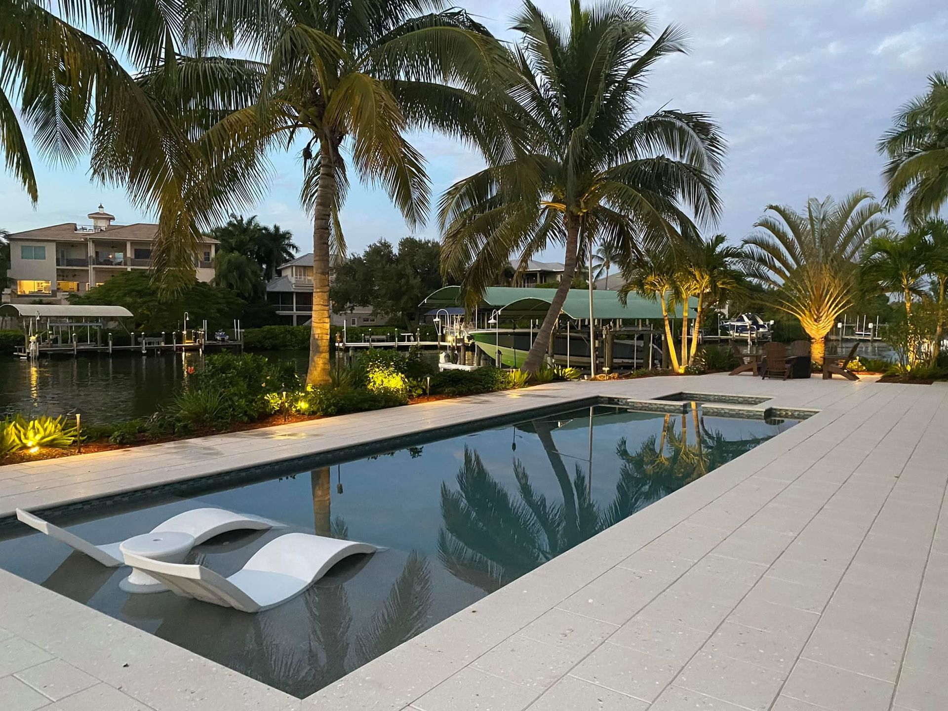Swimming pool with lounge chairs, palm trees, and waterfront homes at dusk.
