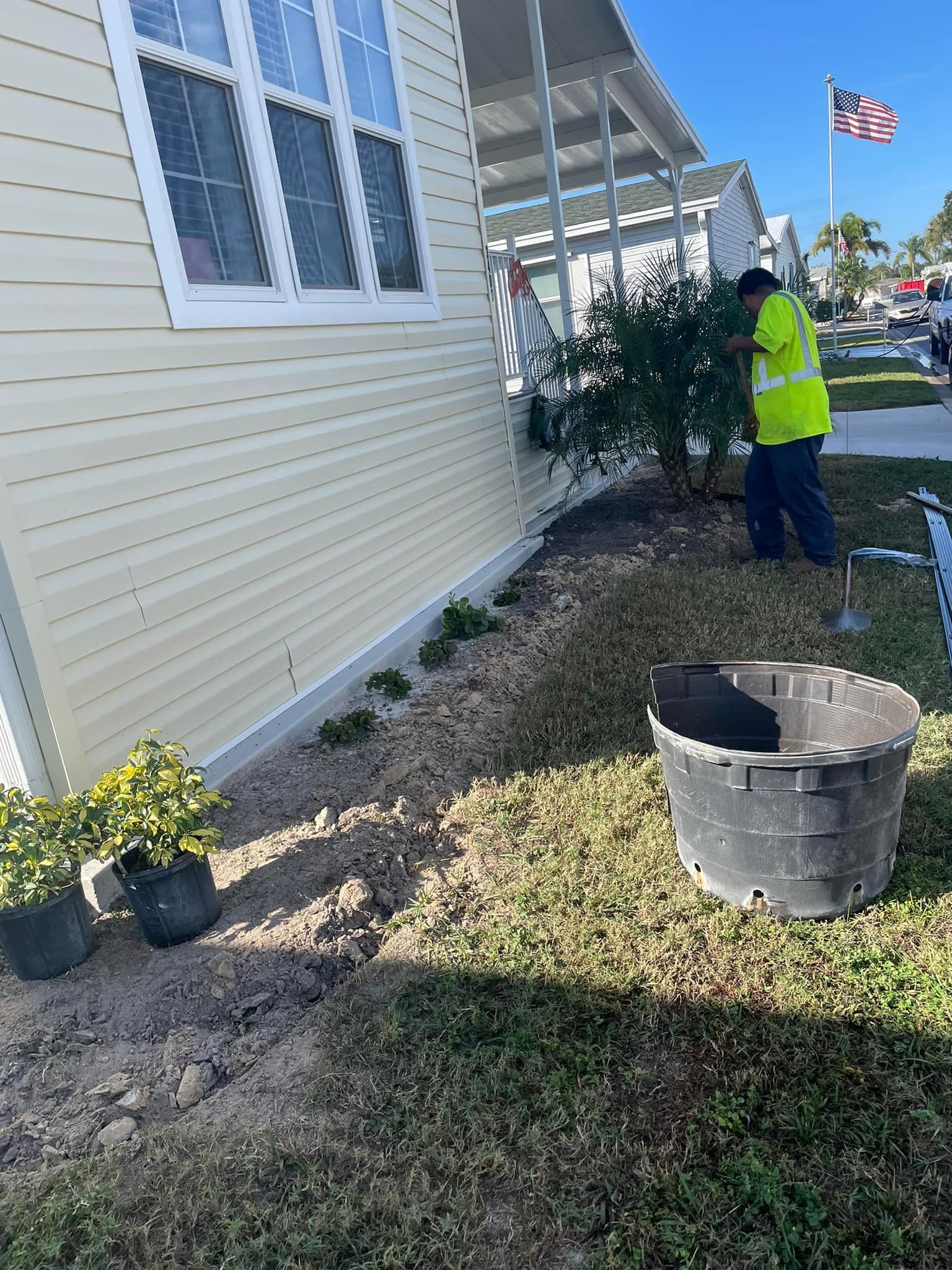Workers planting shrubs next to a light yellow house. Black bucket and potted plants on the grass.