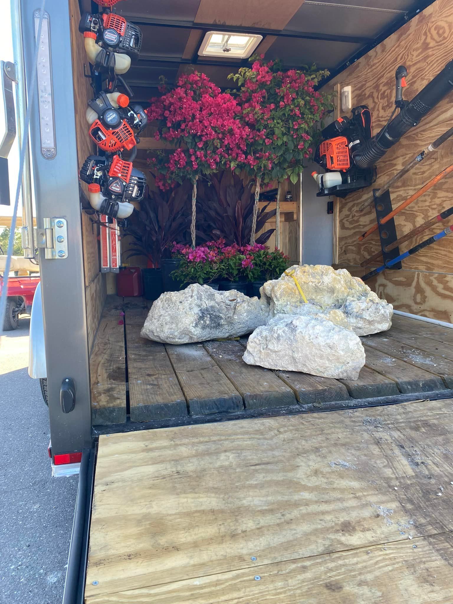 Inside a trailer: rocks, pink flowers, plants, a leaf blower, and orange/black decorations.