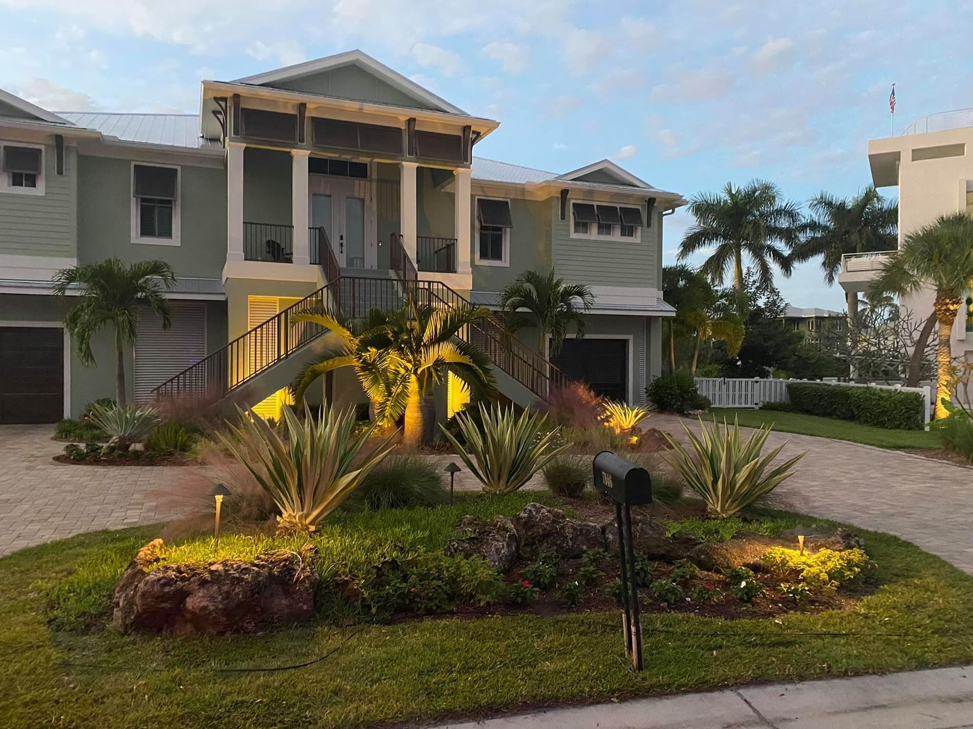 Two-story light green house with gray roof, palms, and landscaped yard. Driveway and mailbox in front.