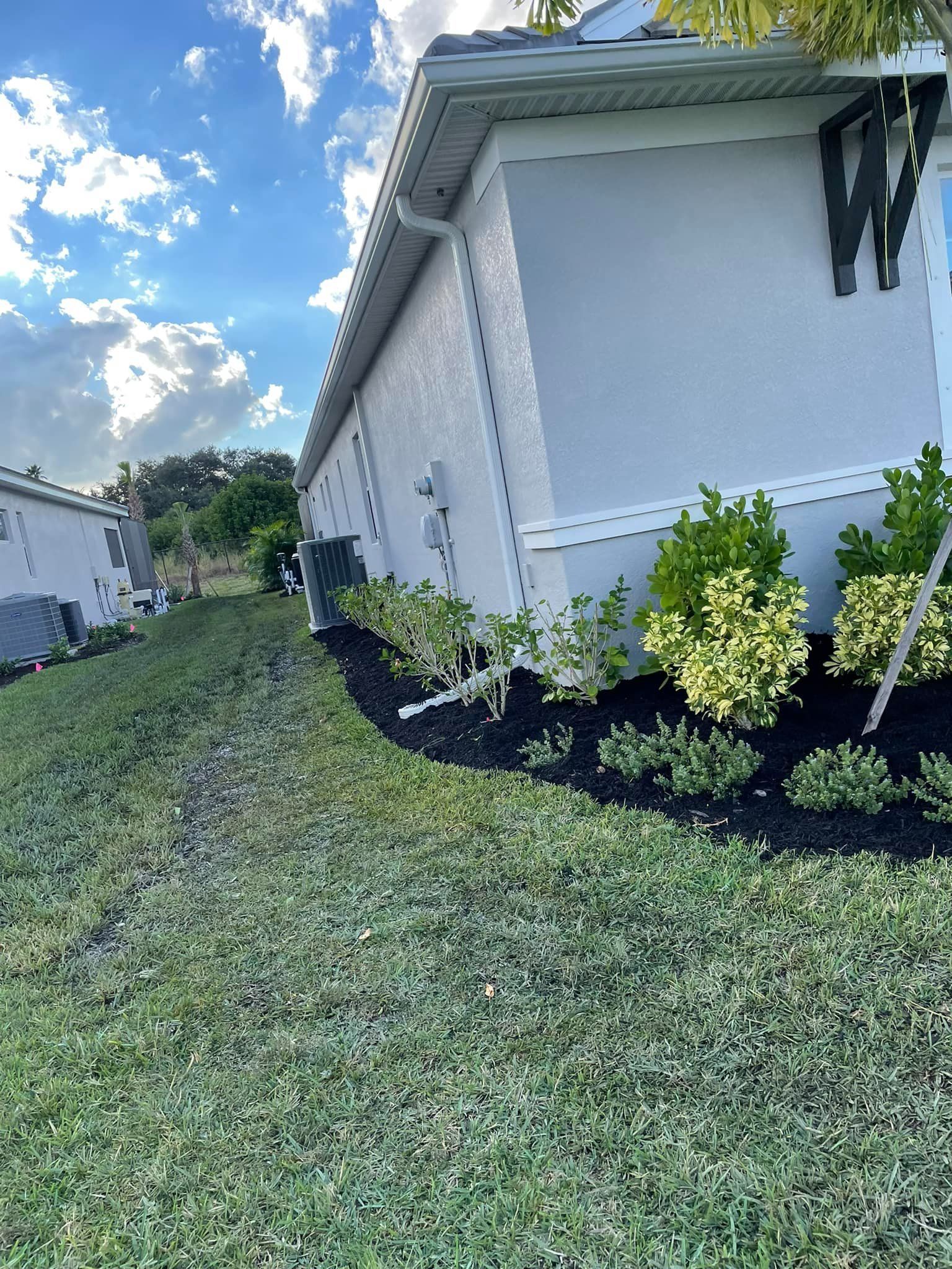 Side view of a light-colored house with green lawn and landscaping under a partly cloudy sky.