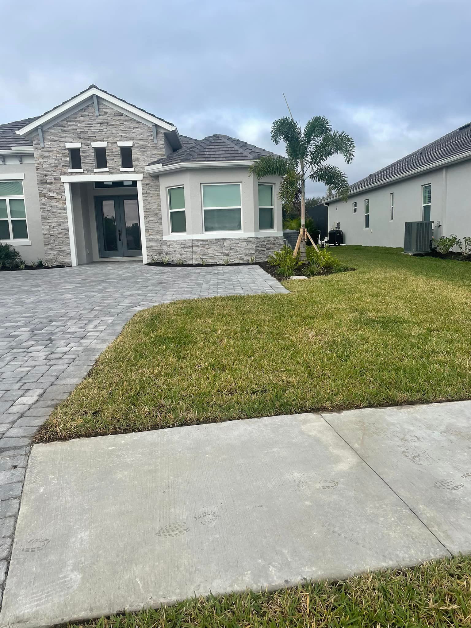 Gray stone facade house with paved driveway and green lawn under a cloudy sky.
