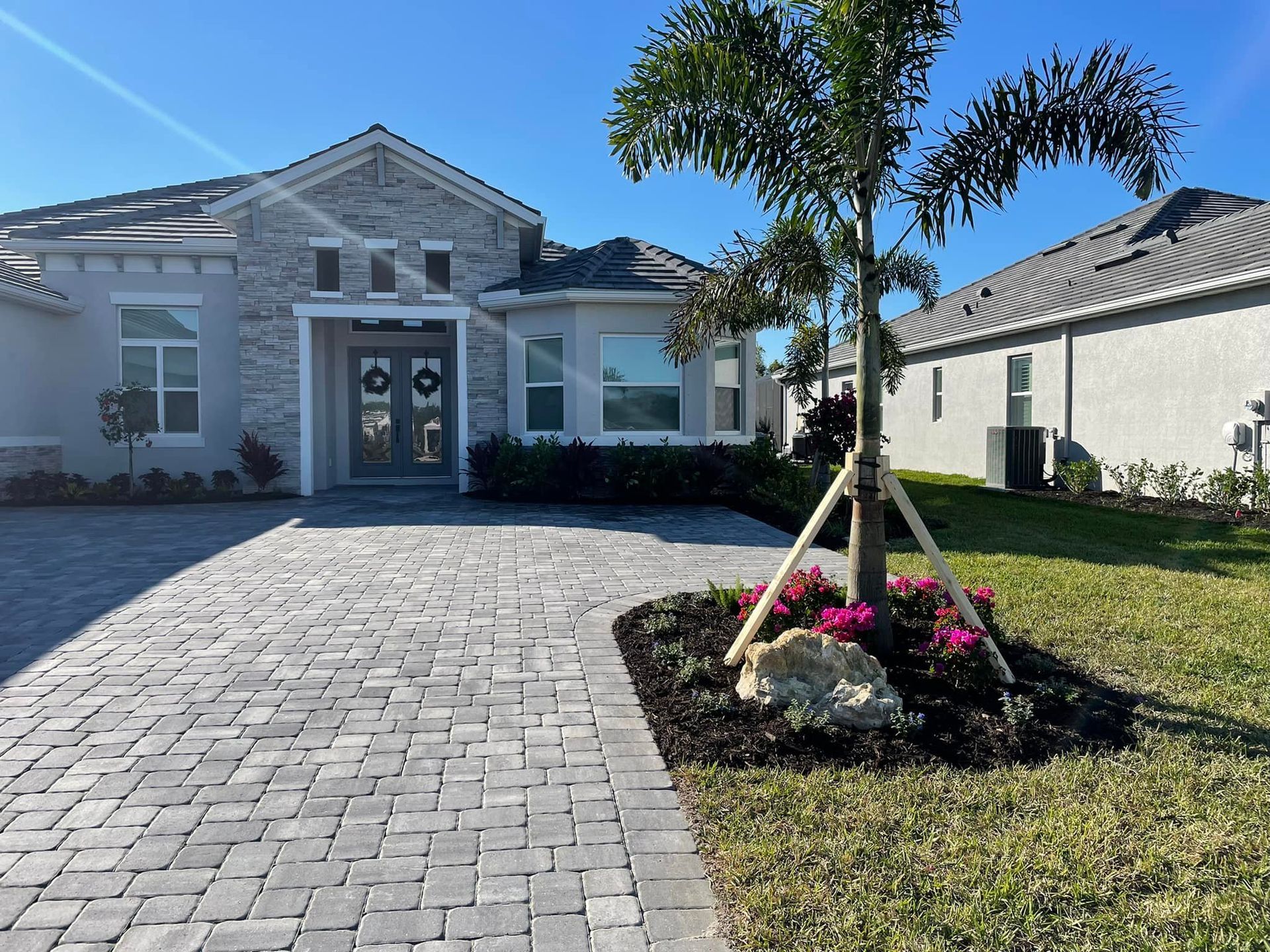 A gray brick house with a paver driveway, front door, and palm tree in the yard under a blue sky.