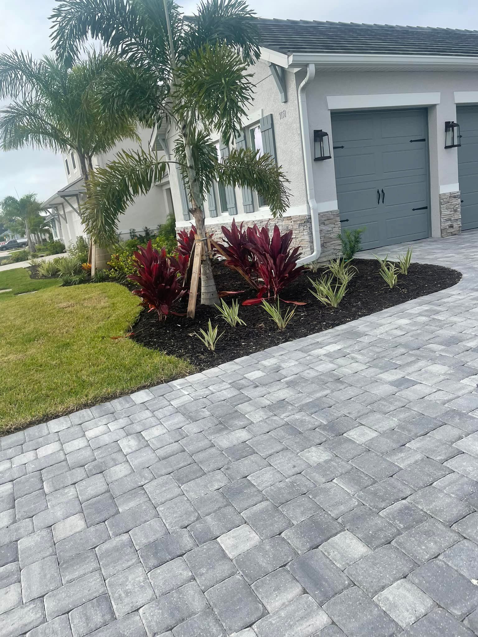 Driveway paved with gray bricks leads to a house with gray garage doors and a landscaped flower bed with red plants.