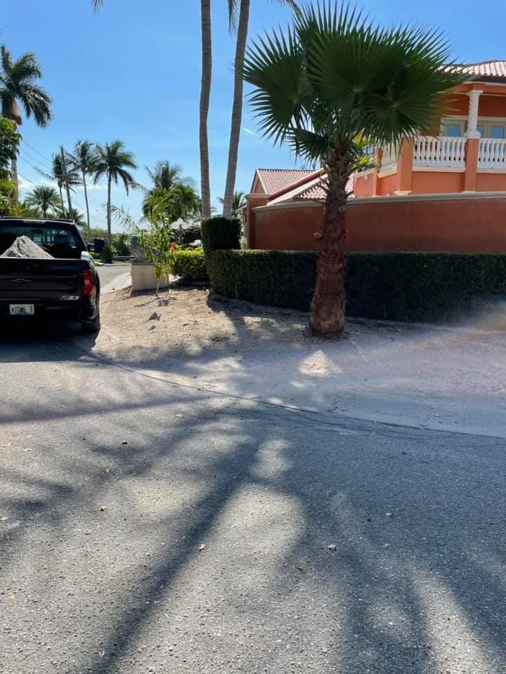 A dirt road with a black truck, palm trees, a hedge, and a peach-colored building on a sunny day.