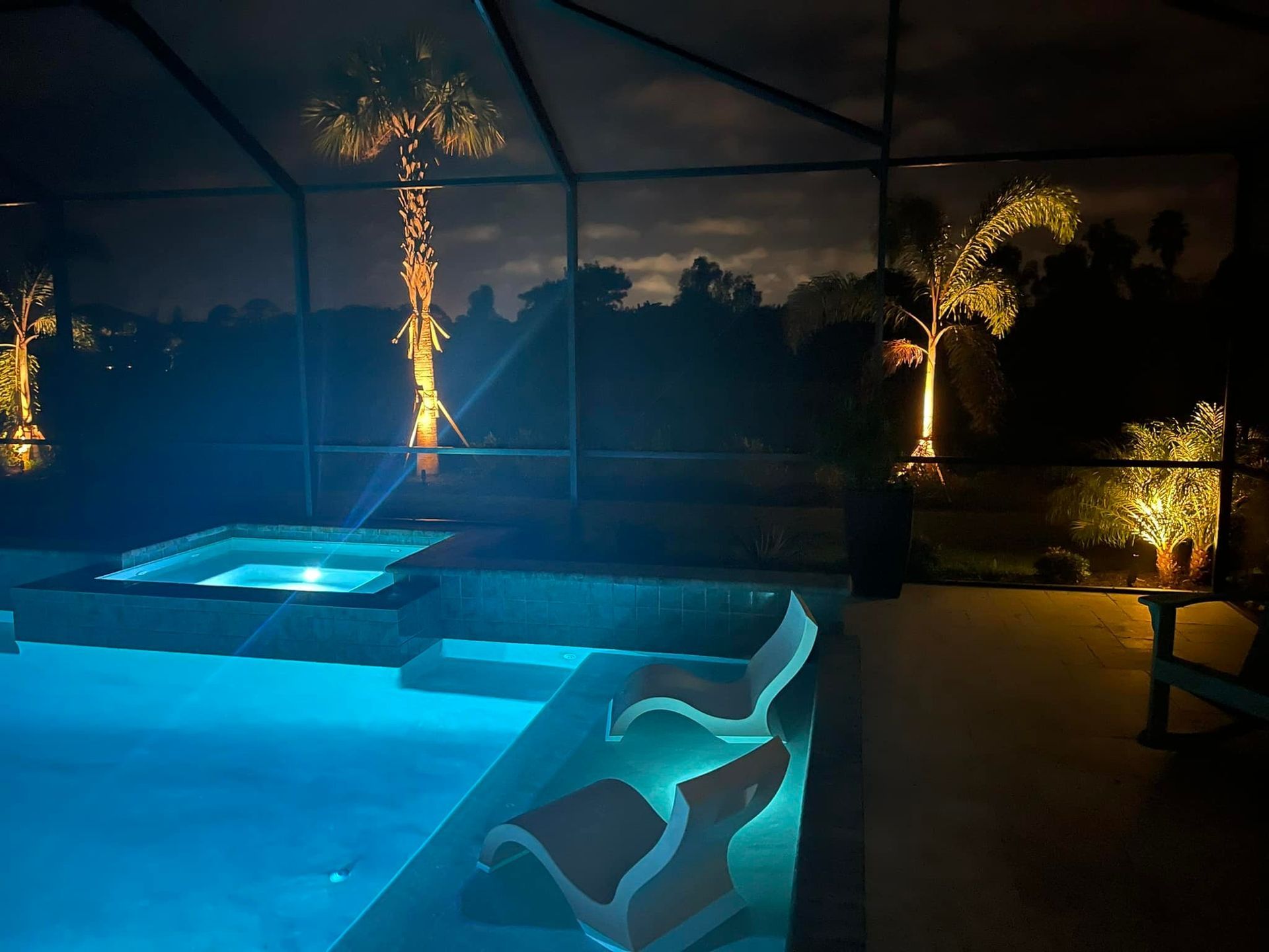 Pool and hot tub at night with underwater and landscape lighting illuminating trees and seating area.