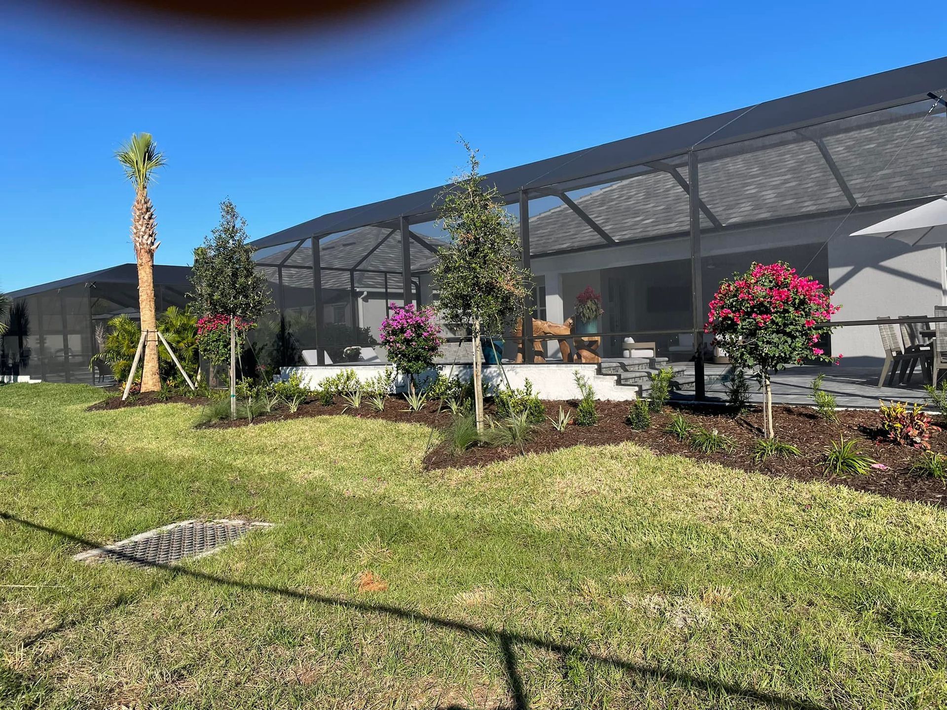 Landscaped backyard with screened patio, flowers, trees, and green lawn under a blue sky.