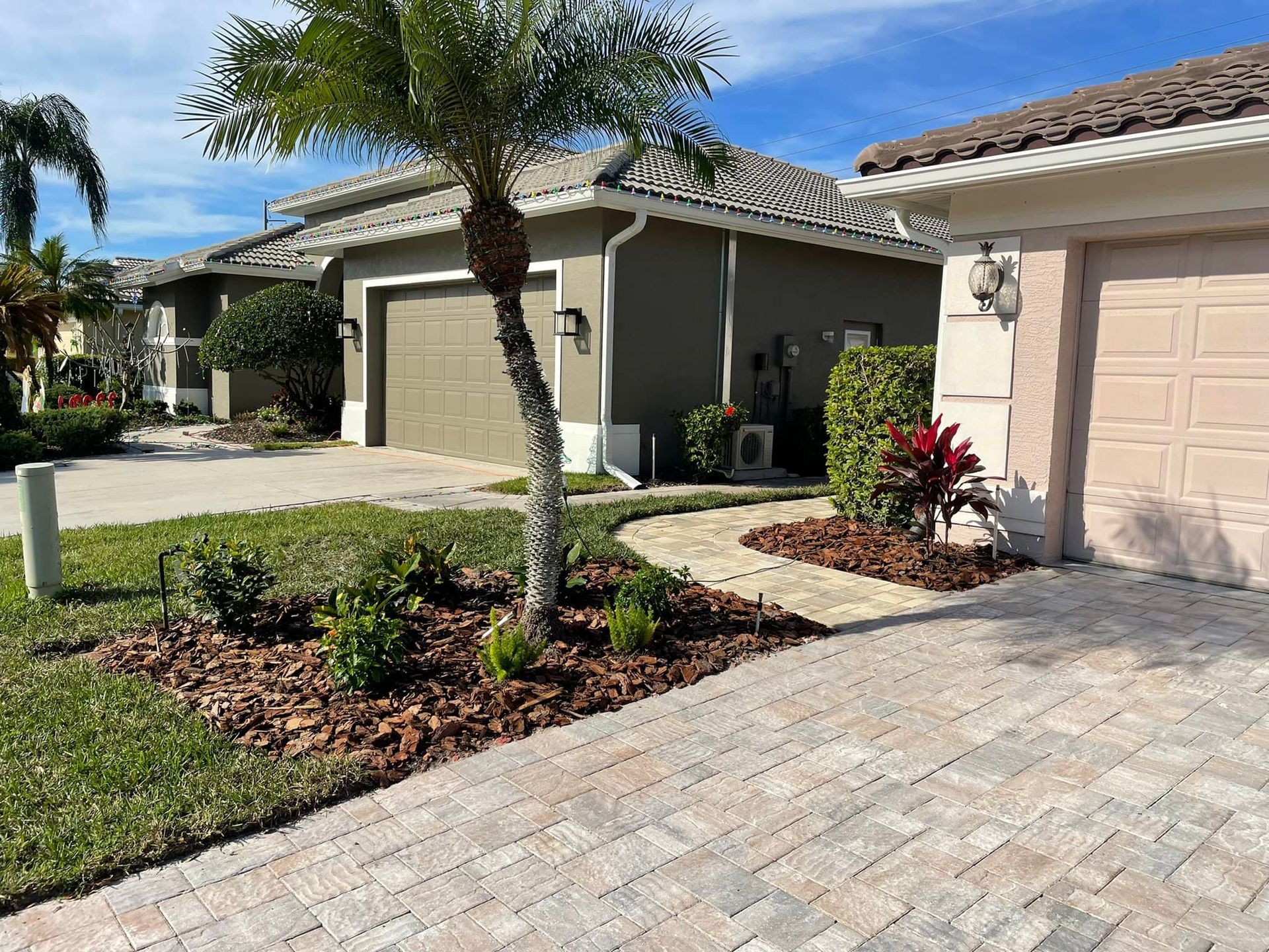Houses with brown garage doors, palm tree, landscaped yard with brick pavers and blue sky.