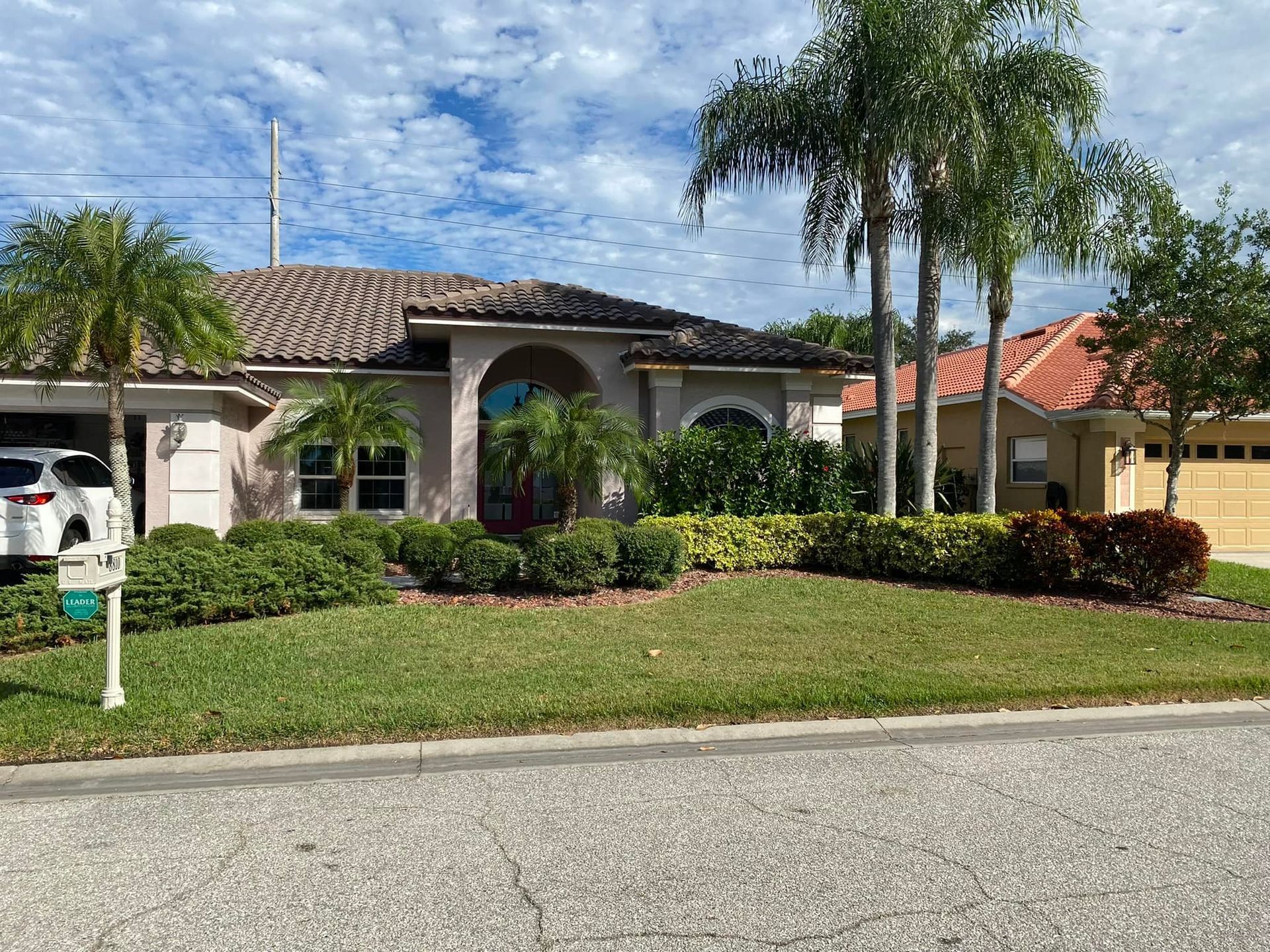 A beige house with a tiled roof, palm trees, and green bushes under a cloudy sky.