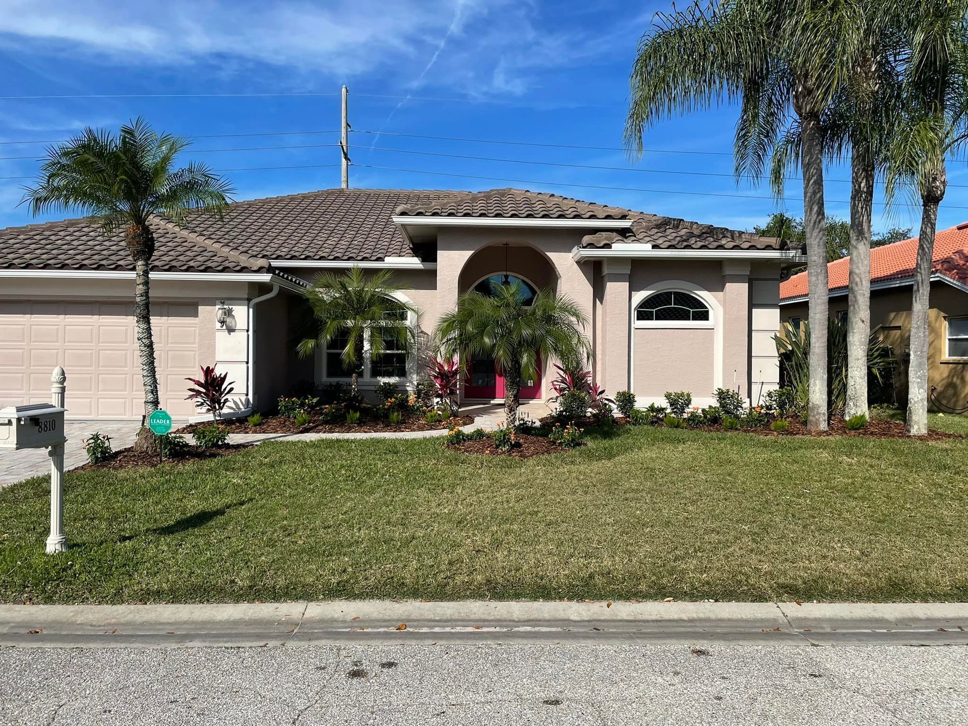 Tan house with tile roof, palm trees, and green lawn against a blue sky.