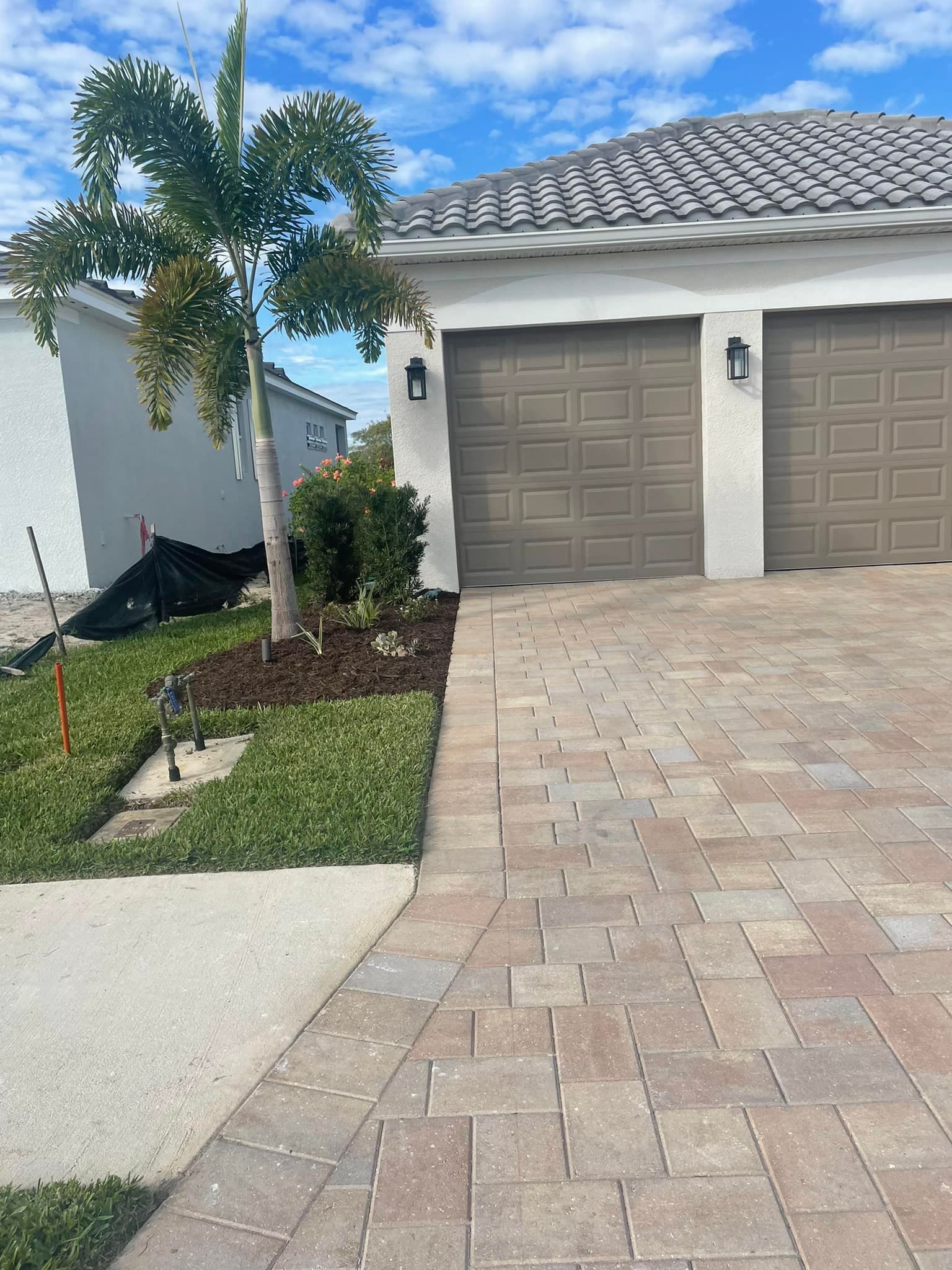 Driveway of a house with a brick paver driveway and a two-car garage. A palm tree is in the landscaped area.