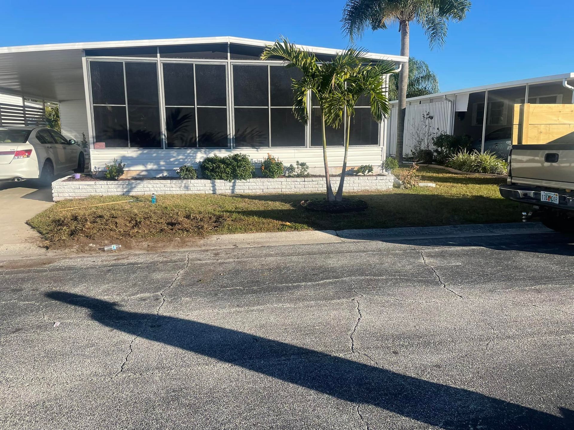 Mobile home with screened-in porch, small palm trees, and car parked in the driveway on a sunny day.