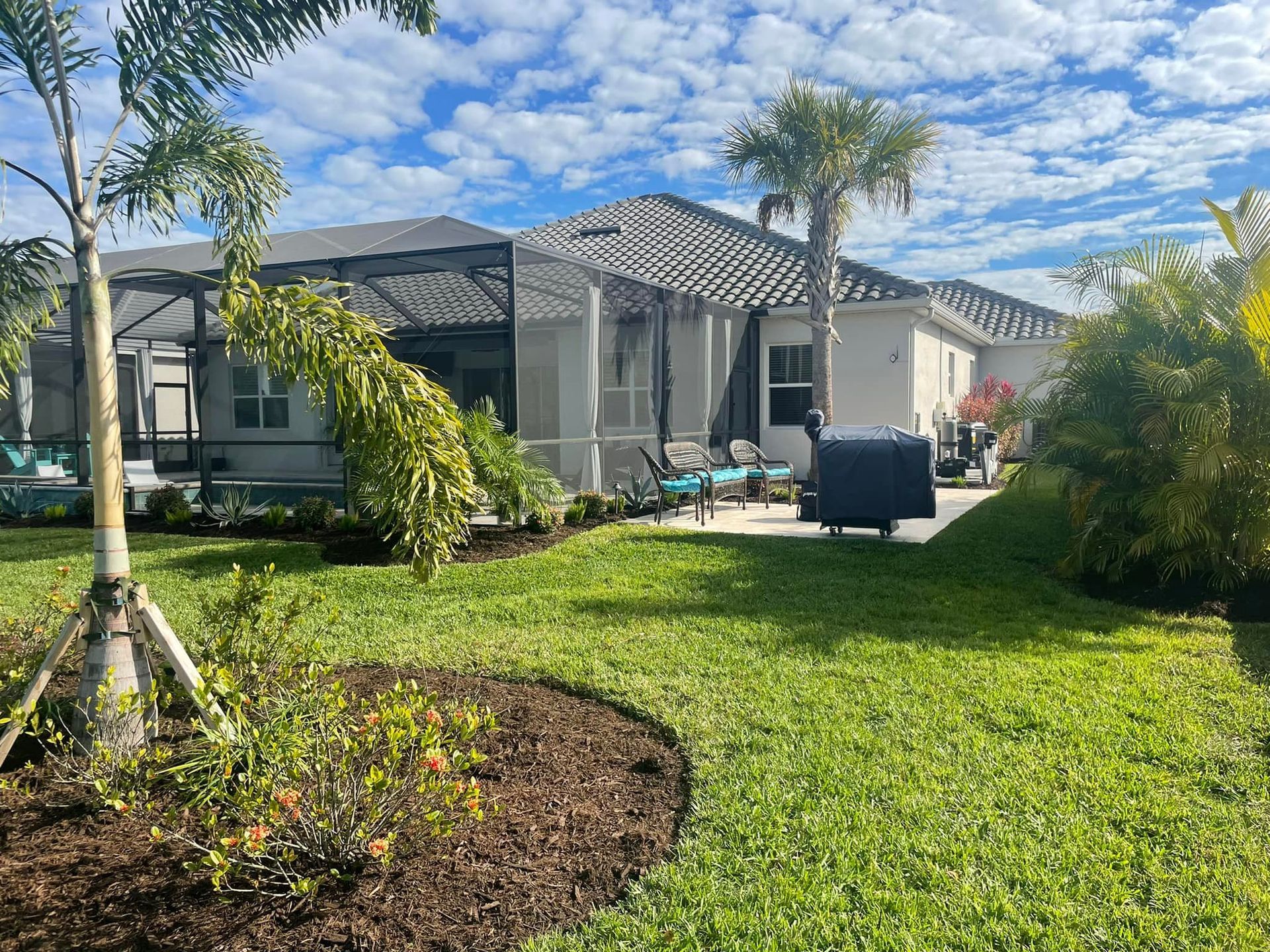 Backyard with green lawn, landscaping, patio, pool cage, and a light-colored house under a blue sky.
