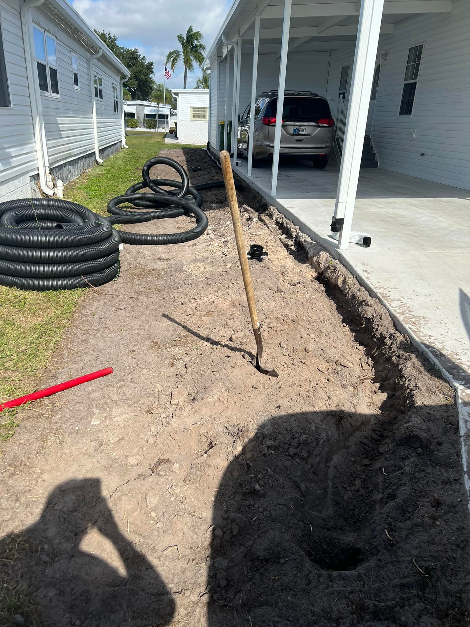 Trench dug alongside a building; black drainage pipes, a hoe, and a car are visible.