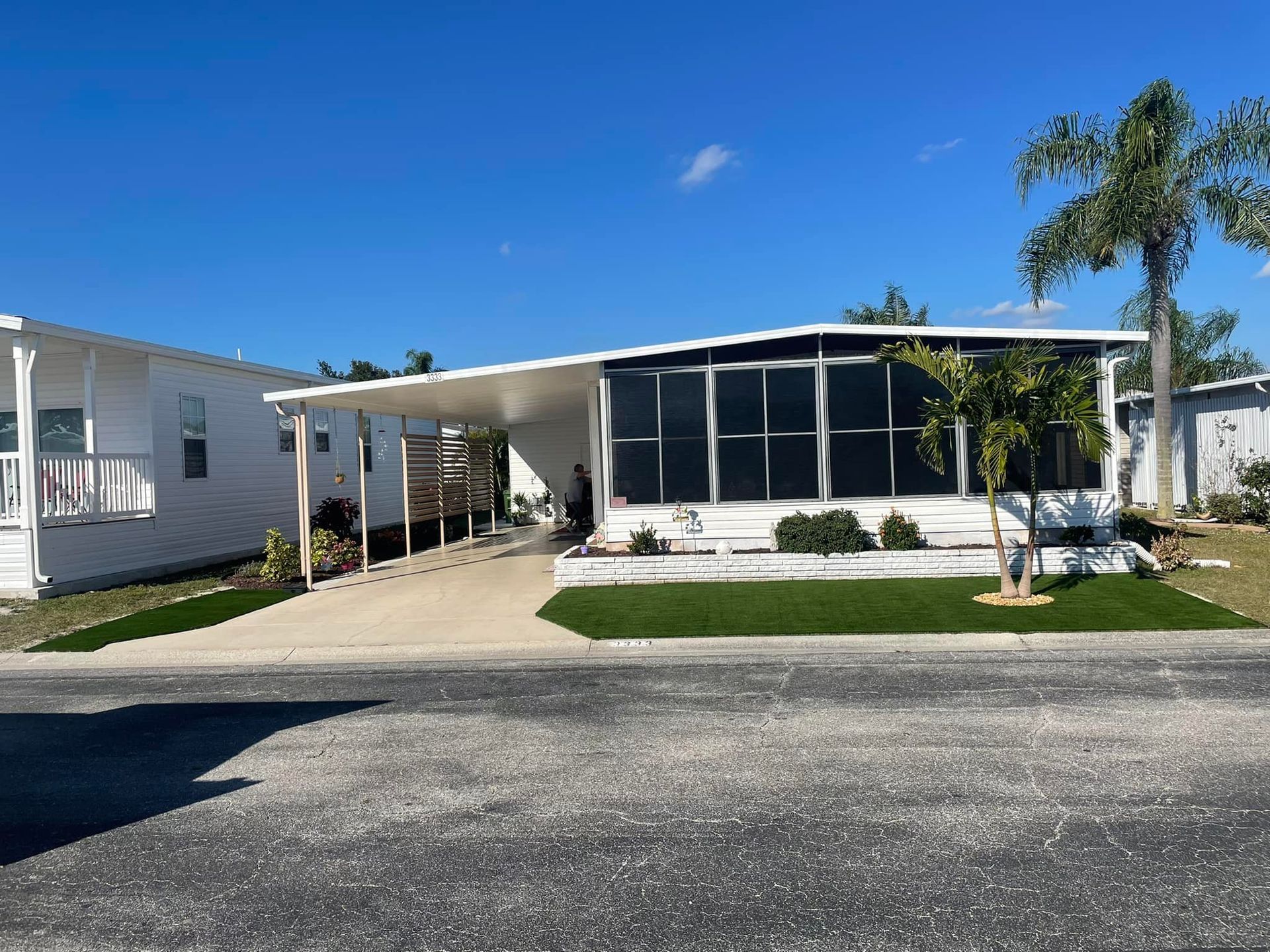 Mobile home with carport, screened porch, and palm tree under a clear blue sky.
