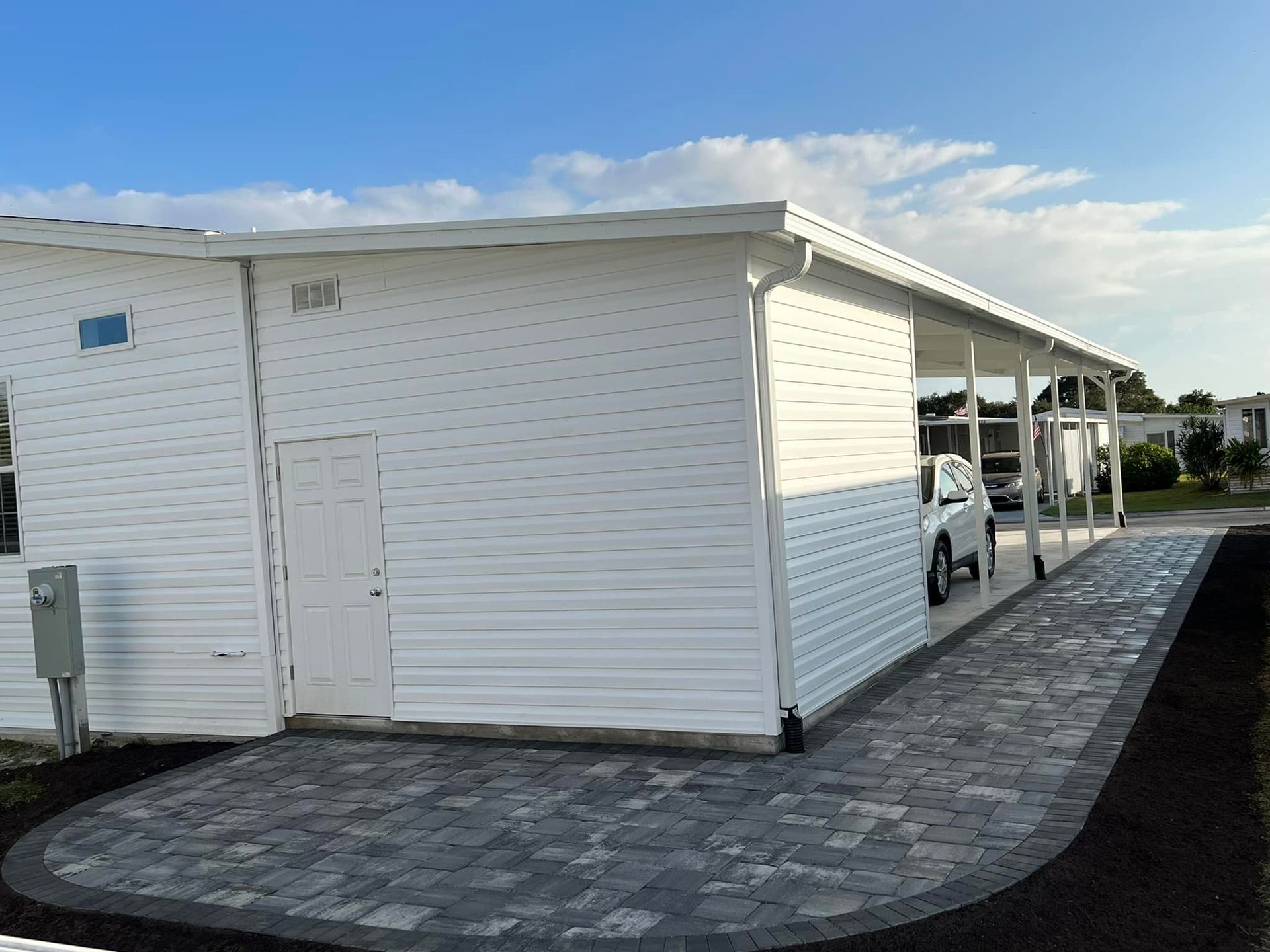 White building with attached carport, gray brick driveway, and white vehicle.